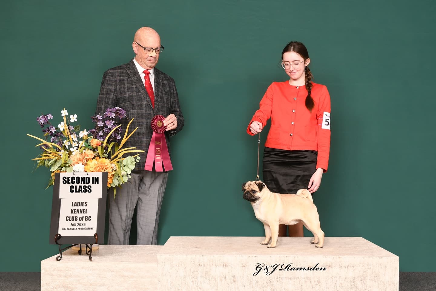 A pug stands on a platform with its handler and a judge holding a second-place ribbon.
