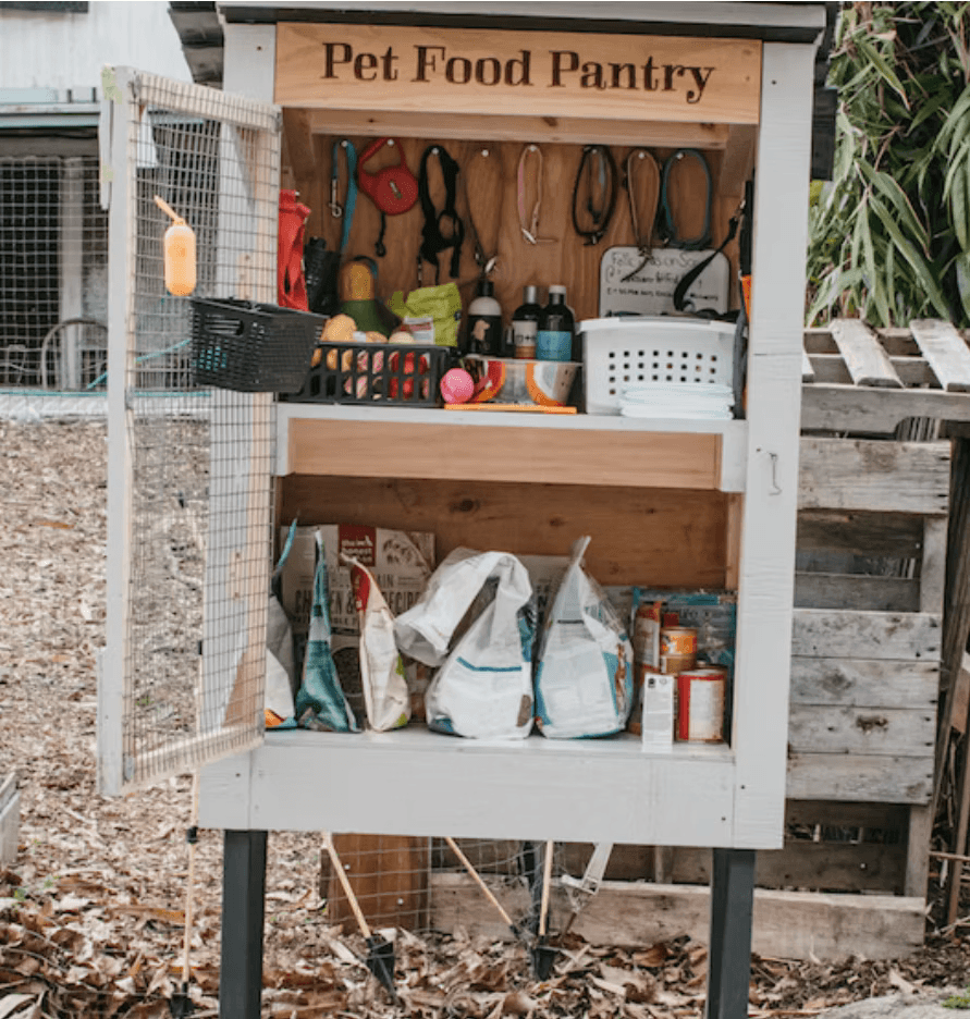 A wooden Pet Food Pantry stocked with leashes, toys, grooming supplies, and bags of food.