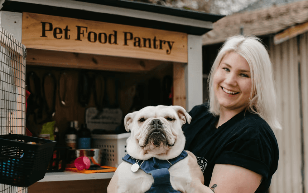 A smiling woman holds a white bulldog in front of a wooden pet food pantry.